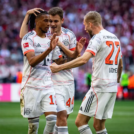 Three football players celebrate on the field, wearing Bayern Munich's white and red away kits. One player with long hair receives an embrace, while two teammates express joy. The background shows a blurred crowd, indicating an energetic match atmosphere.