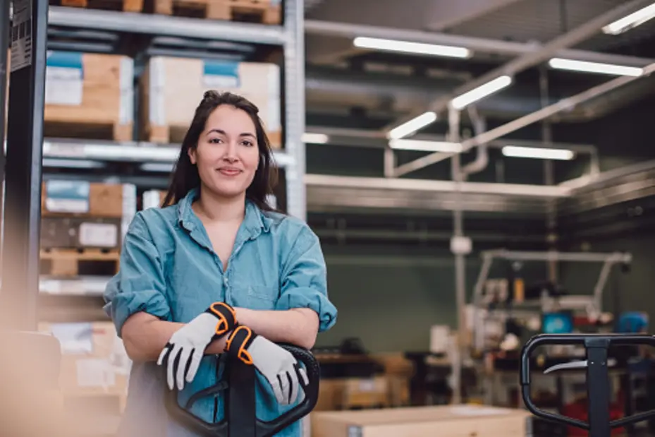 A young woman wearing gloves leans against a pallet jack in a warehouse or storage facility. She has long hair and is smiling, dressed in a denim shirt. The background features shelves filled with wooden boxes and a tidy workspace atmosphere.