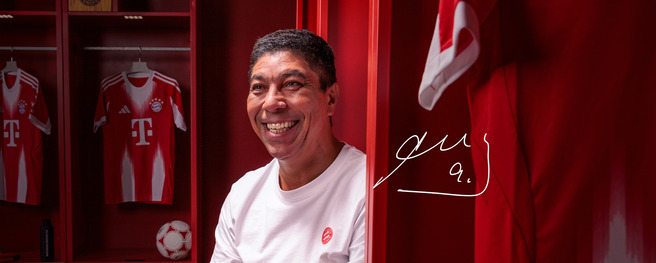 A smiling man in a white shirt sits in a locker room filled with red walls. Behind him are Bayern Munich jerseys hanging on the walls. A football is placed in the locker next to him. The atmosphere is warm and welcoming.