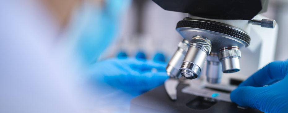 A close-up of a laboratory microscope with a person wearing blue gloves adjusting the focus. The background features laboratory equipment, suggesting a scientific research environment.