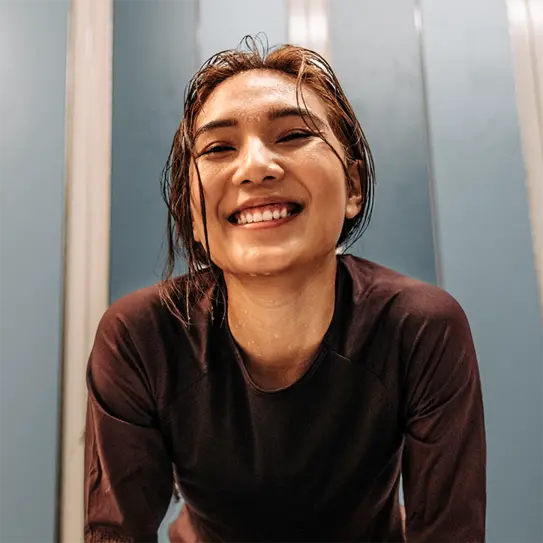A young woman with wet hair smiles broadly at the camera, wearing a dark athletic top. The background features blue panels, suggesting an indoor setting, possibly a gym or sports facility. Her expression conveys happiness and enthusiasm.