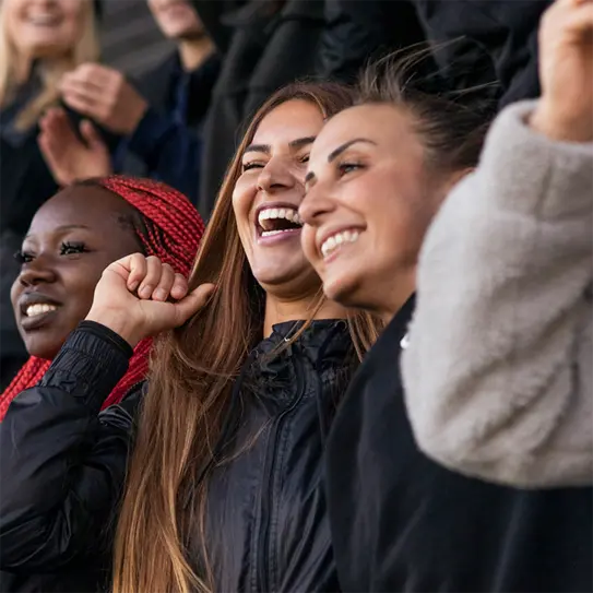 Three young women are joyfully celebrating in a crowd. They have big smiles and are wearing casual jackets. One has long, straight hair, the second has vibrant red braids, and the third has long hair in a ponytail. The atmosphere is lively and positive.