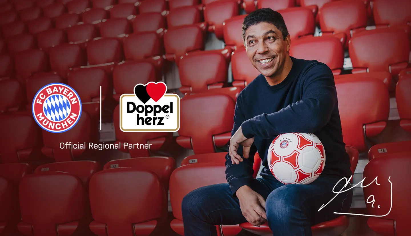 A man smiles while sitting in a stadium seat, holding a soccer ball. The logos of FC Bayern München and Doppelherz are displayed nearby, with the caption "Official Regional Partner" underneath. The background features empty red stadium seats.