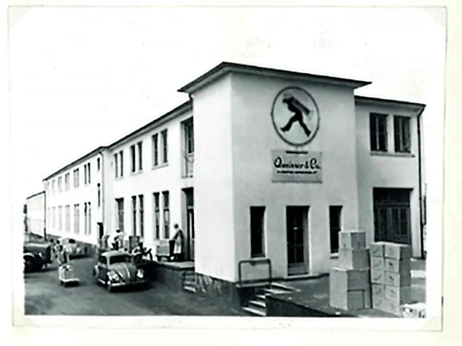 A black and white historical photograph of a large building with a prominent logo featuring a figure in motion. People are seen near the entrance and parked cars are visible. Stacks of boxes are positioned in the foreground, suggesting an operational or commercial setting.