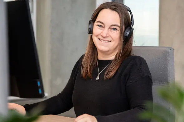 A woman with long brown hair smiles while sitting at a desk wearing headphones. She is in front of a computer monitor with a blurred background, suggesting a professional work environment.