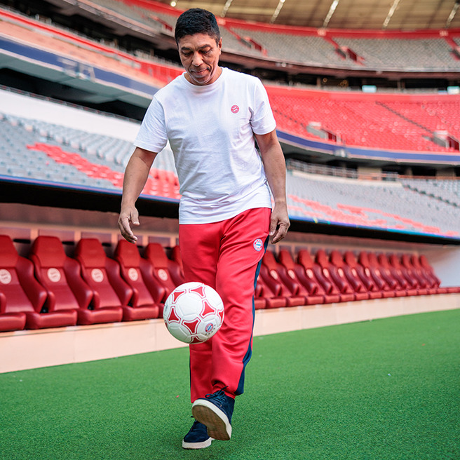 A person in a white shirt and red athletic pants is skillfully juggling a soccer ball on a green field inside a stadium. Empty red seating is visible in the background, showcasing the venue's sports atmosphere.