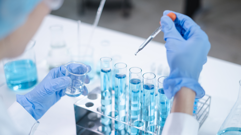 A scientist wearing gloves and a lab coat is holding a beaker with blue liquid in one hand and a pipette in the other, preparing to add drops to a rack of test tubes filled with the same blue liquid.