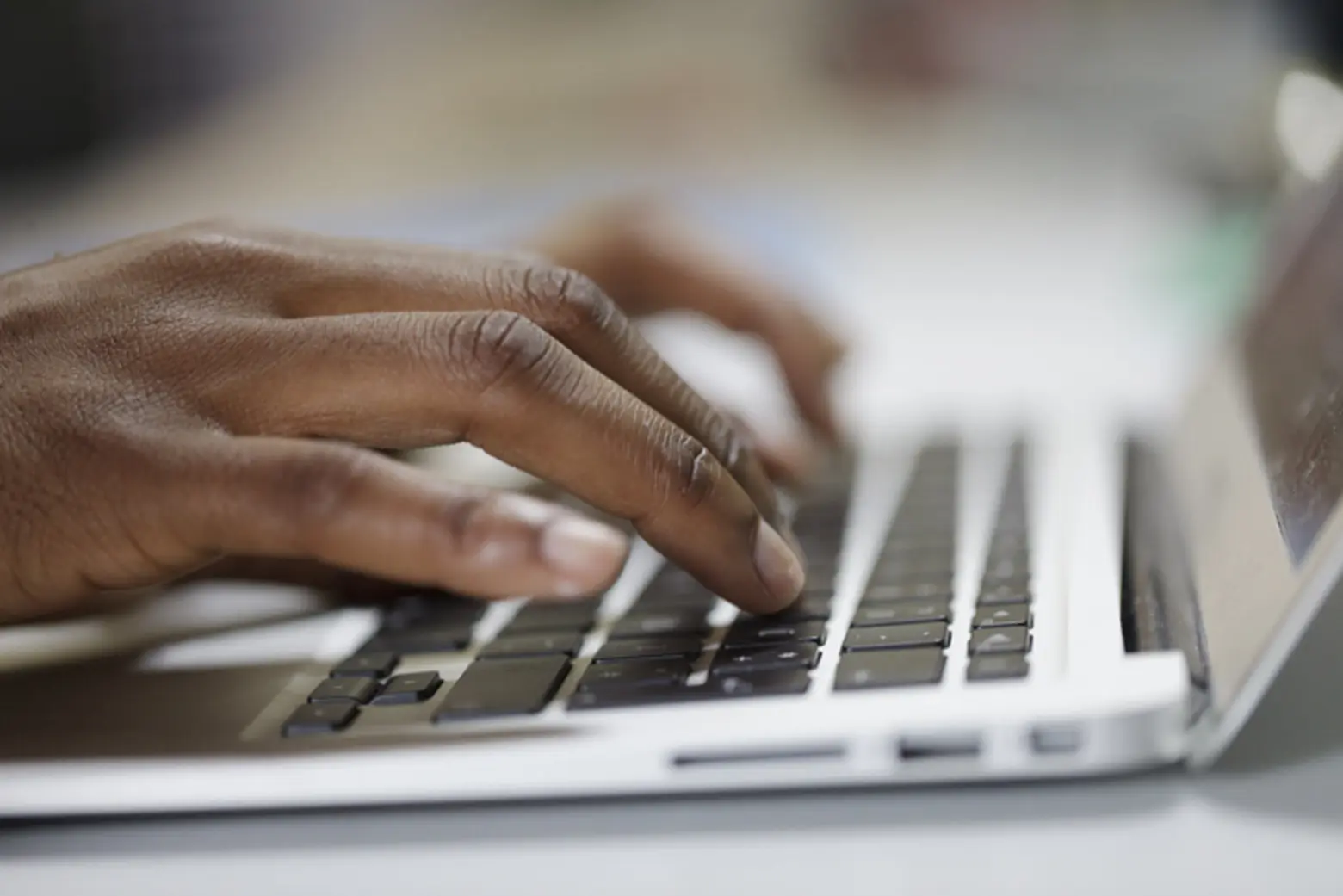 A close-up of a hand resting on a laptop keyboard, with fingers poised over the keys as if typing. The background is blurred, emphasizing the action of using the computer.