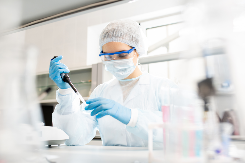 A scientist wearing a lab coat, gloves, goggles, and a face mask is using a pipette to transfer liquid in a laboratory setting. The background features laboratory equipment and glassware.