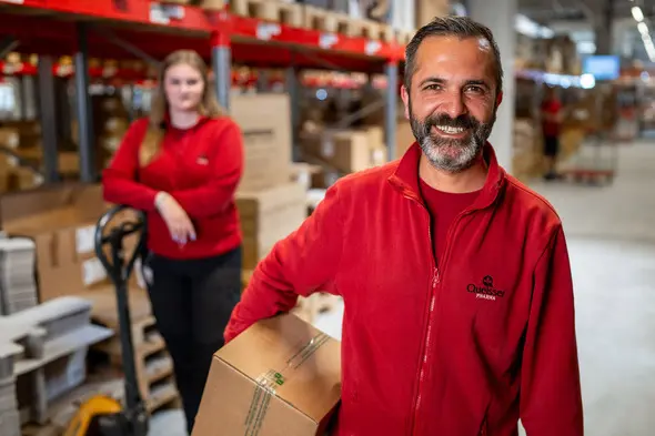 A smiling man in a red uniform holds a cardboard box in a warehouse, with shelves stocked in the background. A woman in a red shirt stands behind him, leaning on a pallet jack, both appearing engaged in their work environment.
