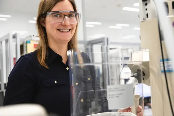 A smiling woman wearing safety glasses stands next to a laboratory machine. She is dressed in a black shirt and appears to be in a clean, modern lab environment. Various lab equipment can be seen in the background.