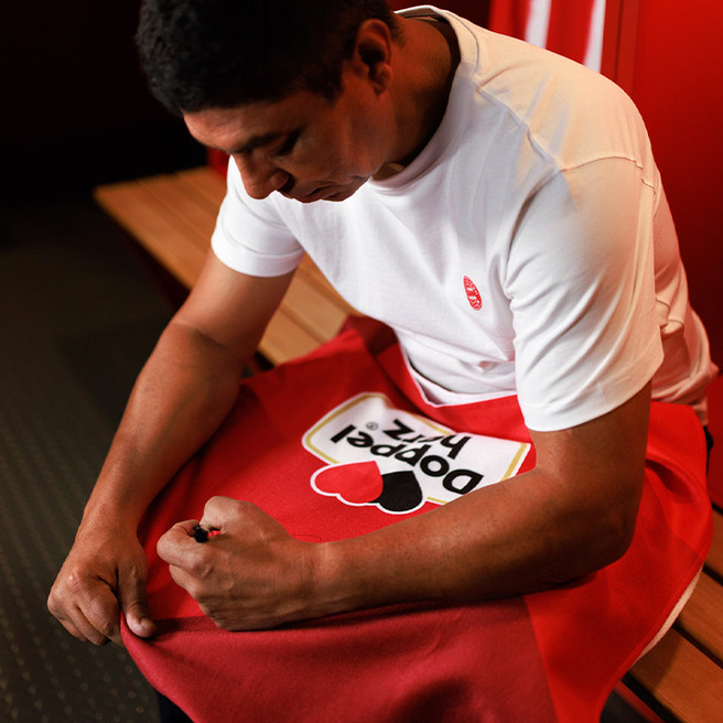 A man sits on a wooden bench in a locker room, focusing on adjusting a red towel with a graphic that reads "Popper Pix." He is wearing a white shirt and has a determined expression as he works on the towel.