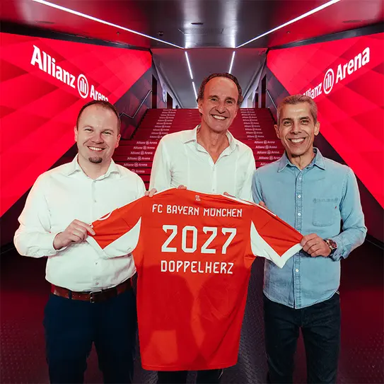 Three men are standing in front of a brightly lit hallway at Allianz Arena, each holding a red FC Bayern Munich jersey featuring the year "2027" and the word "DOPPELHERZ." The vibrant background enhances the celebratory atmosphere.
