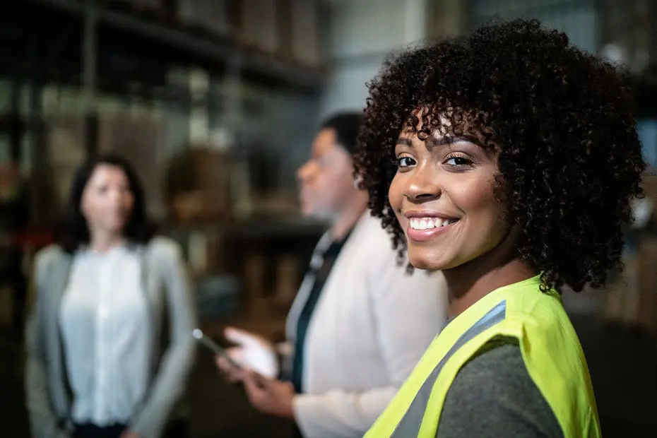 A smiling woman with curly hair, wearing a yellow safety vest, stands in a warehouse setting. Two other women are slightly blurred in the background, engaged in conversation. The scene suggests a professional environment focused on teamwork and communication.