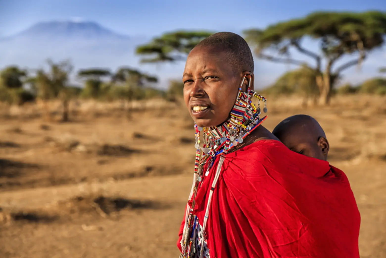 A woman wearing traditional Maasai attire, including a vibrant red shuka and intricate beadwork, stands in a rural landscape. A child is carried on her back, and the distant silhouette of a mountain is visible against a clear blue sky.